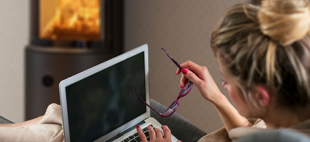 Woman with hair top sits in front of a black Scan 67 wood stove with a PC in her lap and glasses in her hand