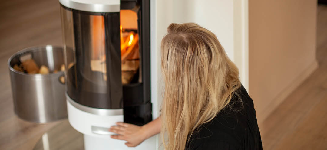 A blonde woman sits next to a white round Scan 83-2 wood stove and pushes the door shut