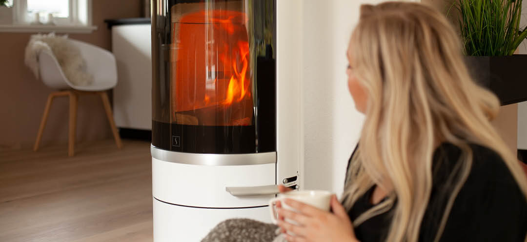 Blond woman sits by a white Scan 83-2 wood stove and drinks tea