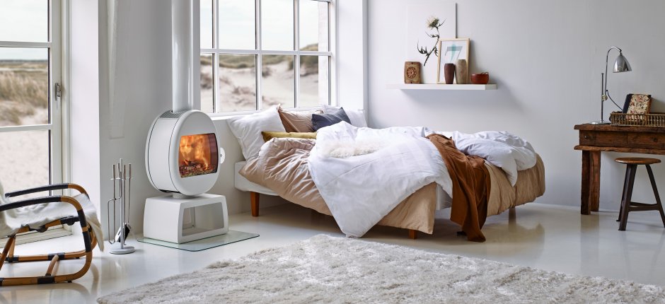 White wood-burning stove Scan 66 in a white one-room cabin next to a bed and desk in wood