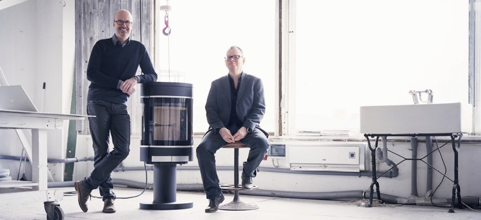 Two middle-aged men in a bright factory room, one leaning against a black fireplace Scan 68 and the other sitting on a stool